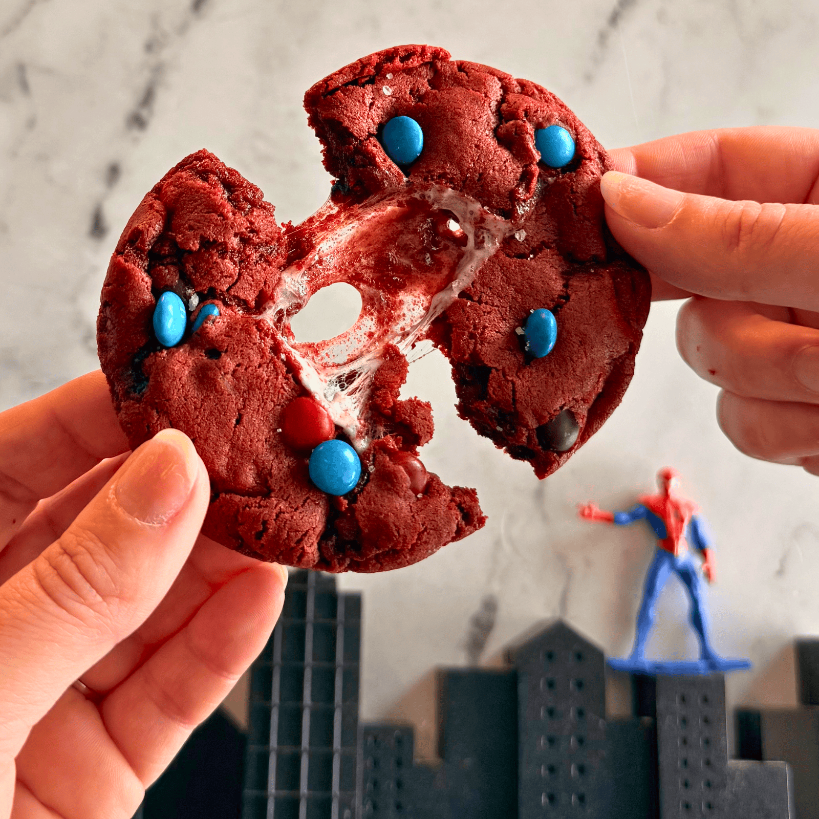 a hand holding a Spider-Man Cookie pulling two halves apart, with marshmallow pulling between, and a skyline and small figurine in the background.