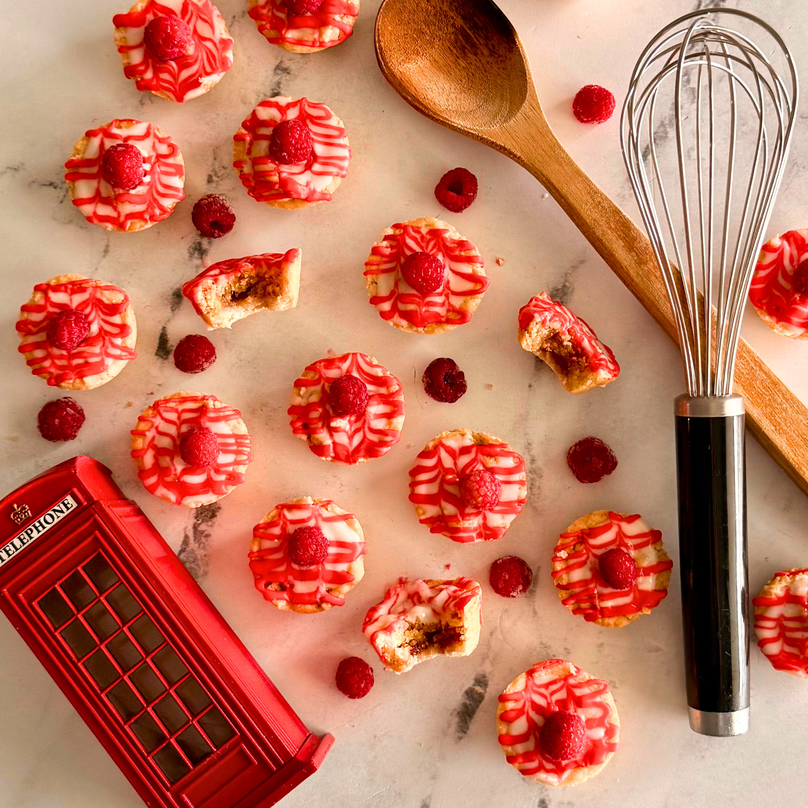 about a dozen bakewell cookie cups on a marble surface, with a wooden spoon, a whisk, and a british phone box nearby and raspberries in between.